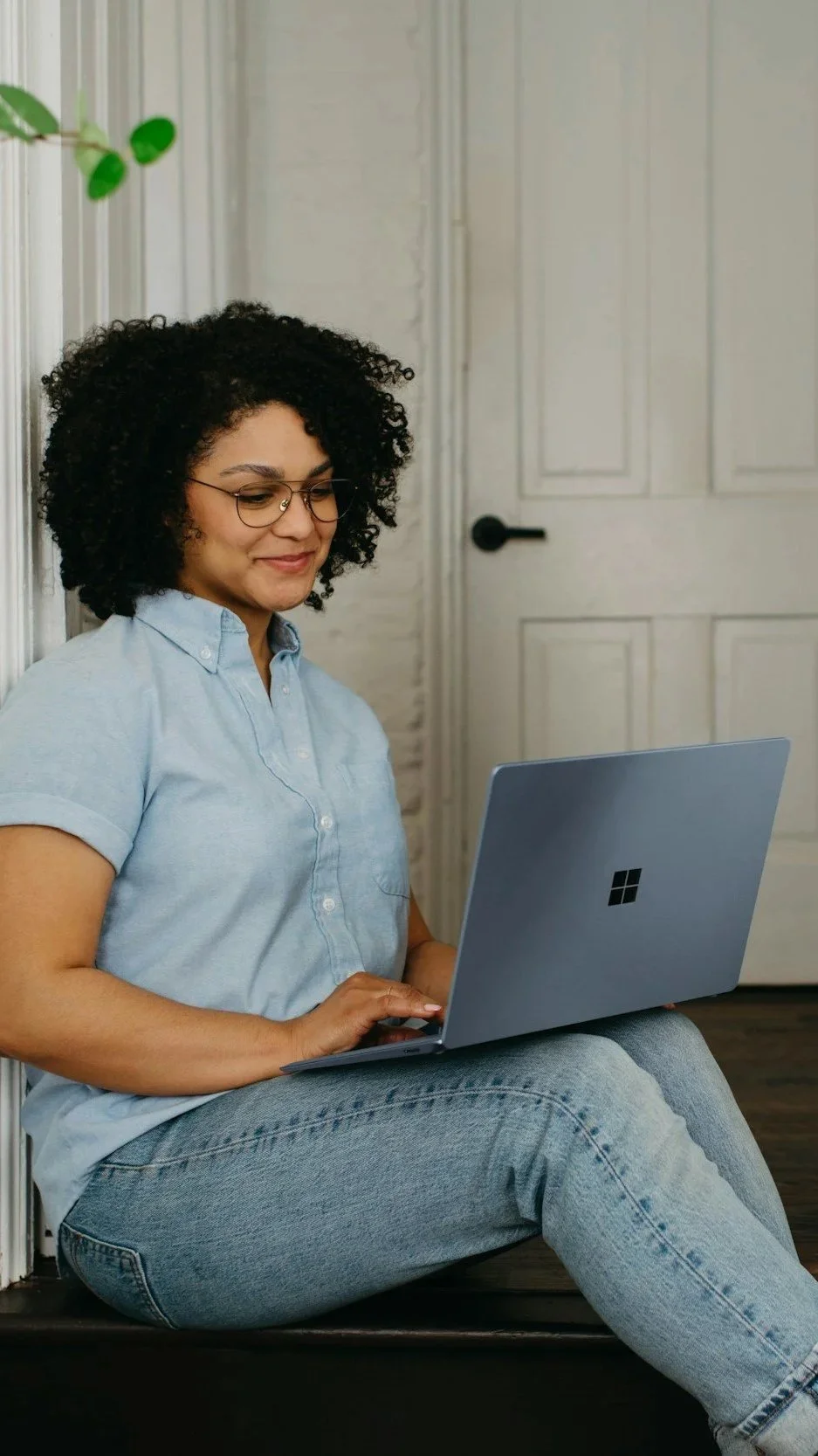 A young woman with curly black hair, glasses, wearing a light blue shirt and jeans, sitting on the floor, looking at a silver Microsoft Surface laptop.