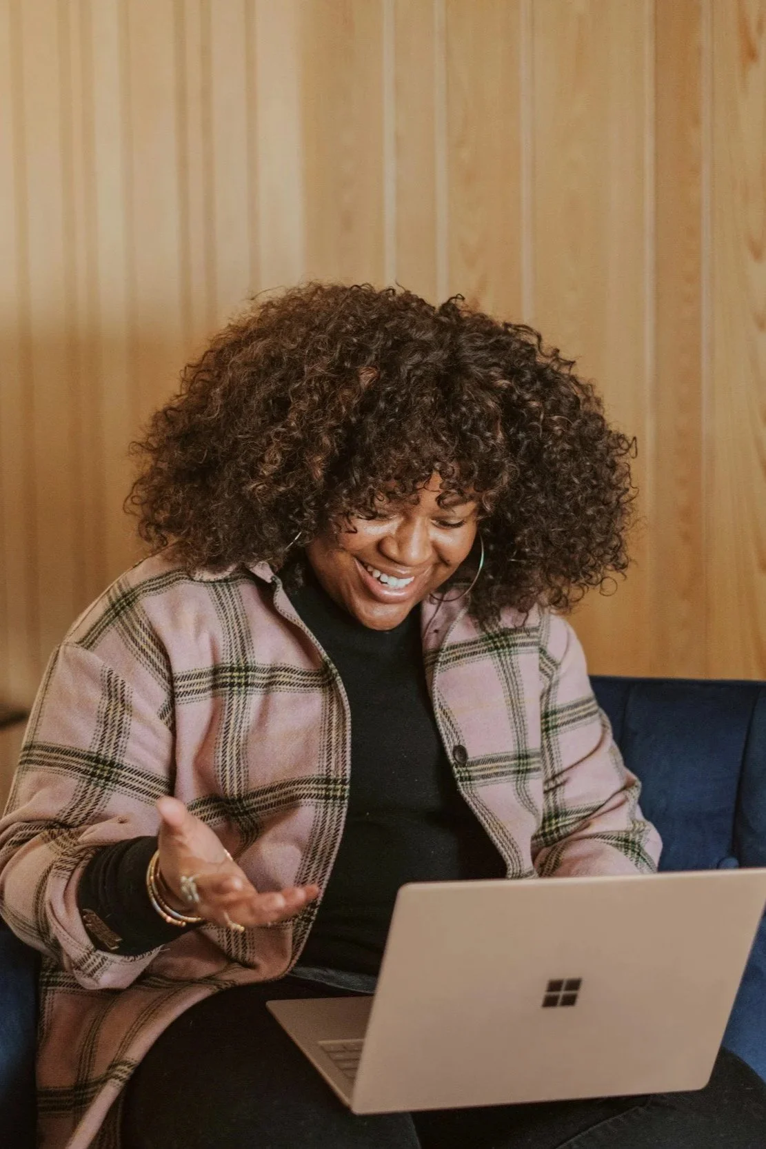 A woman with curly hair, wearing a plaid jacket, sitting on a couch in front of a laptop, smiling and engaging in a video call or looking at something on the screen.
