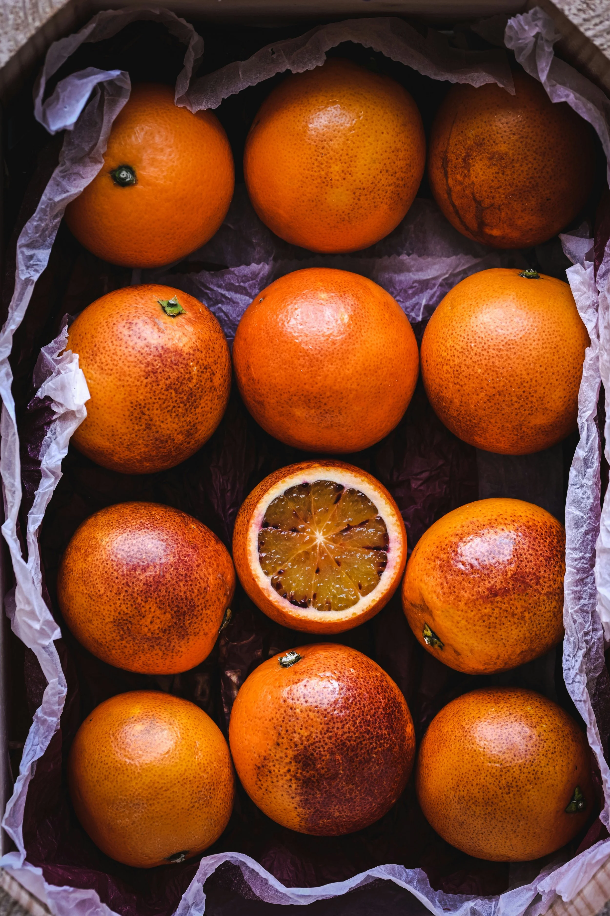 A box containing twelve blood oranges, with one cut in half revealing its inner flesh.
