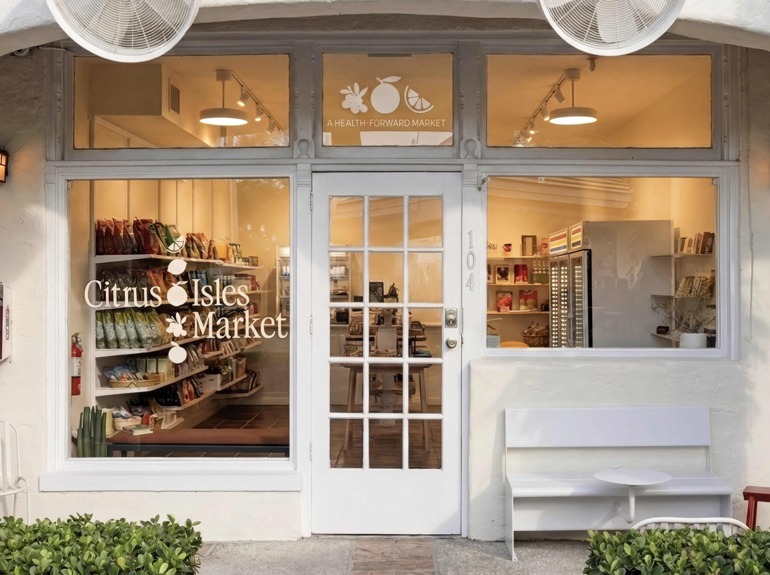 Exterior of Citrus Isles Market, a small shop with large front windows displaying the store's name, with some greenery outside and a white bench seating area.