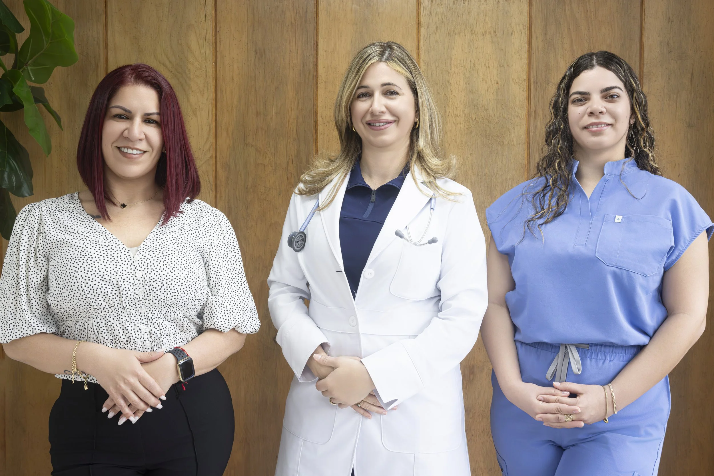 Three women, possibly healthcare professionals, standing together against a wooden background. The woman in the center is wearing a white doctor's coat with a stethoscope around her neck, flanked by two women in blue medical scrubs and casual clothing.