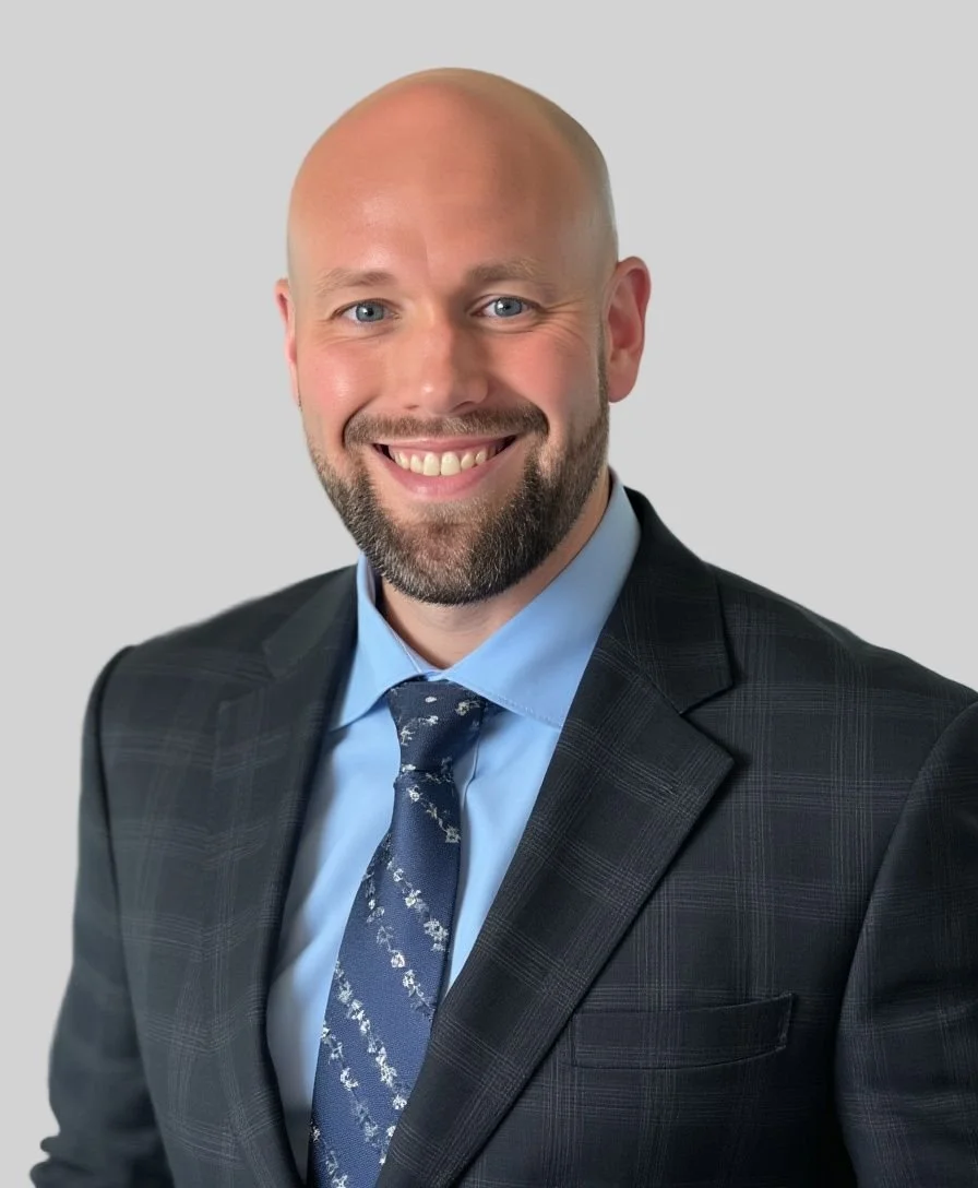 A smiling man with a beard in a suit and tie, against a plain light gray background.