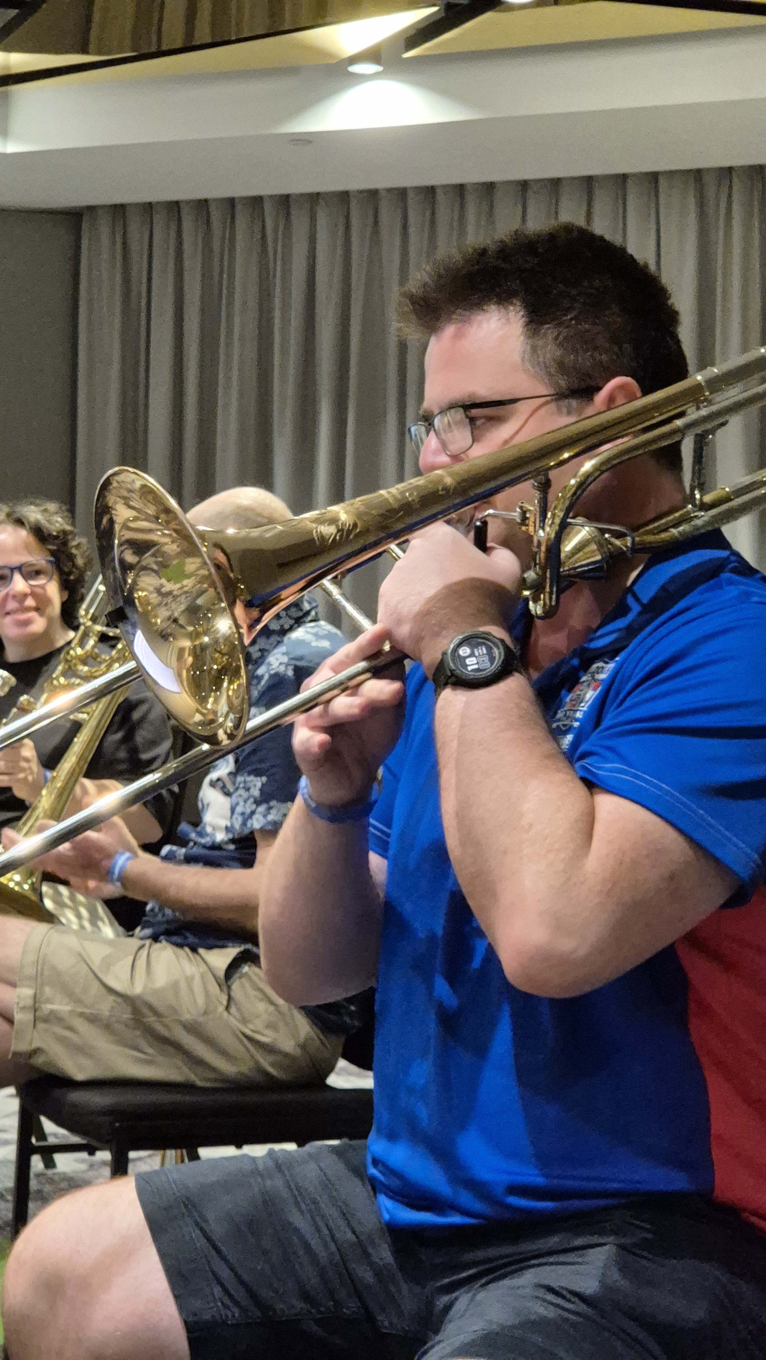 A group of people playing brass instruments, including a man in a blue shirt and glasses, sitting on chairs in a room with gray curtains and ceiling lights.