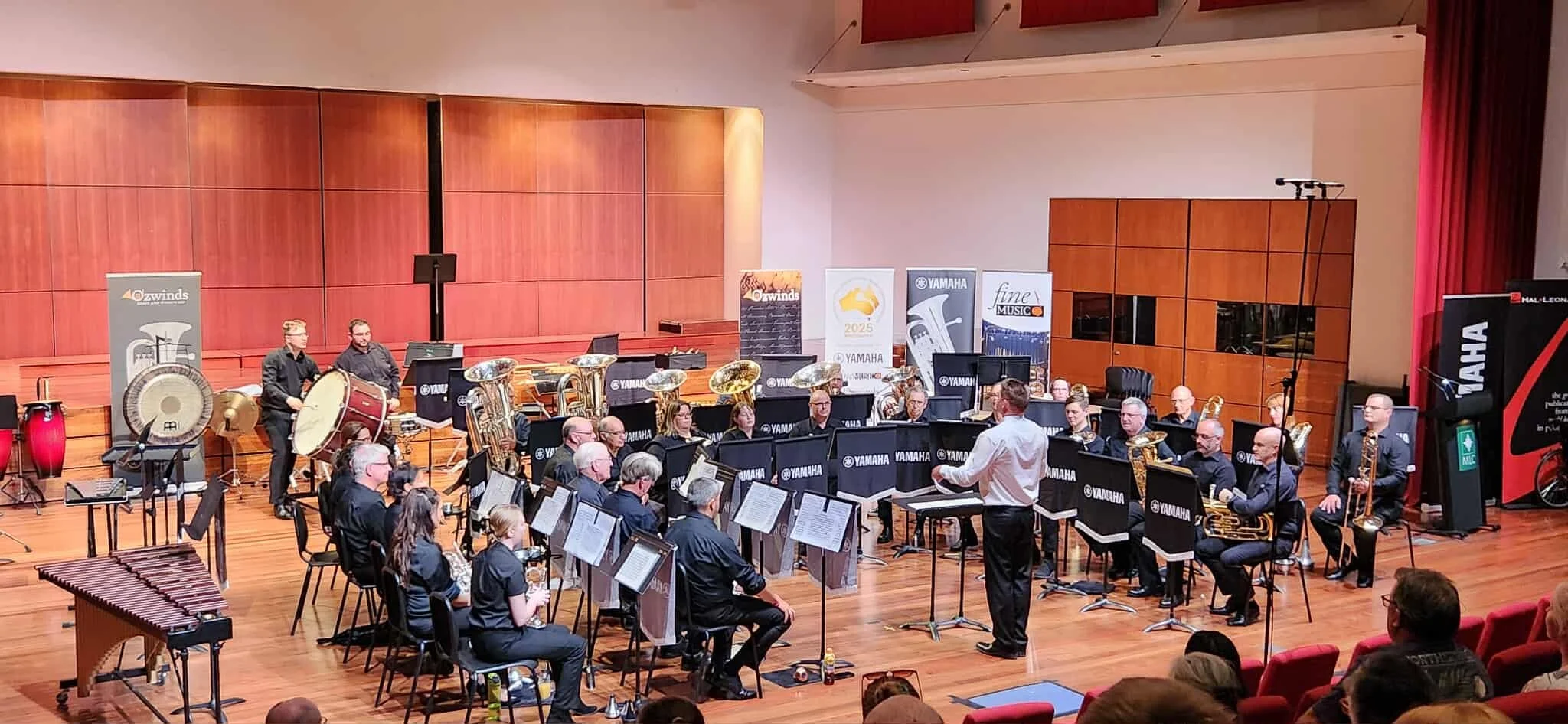 An orchestra performing on stage in a concert hall with wooden panels and banners, conductor leading the musicians, and audience in red chairs.