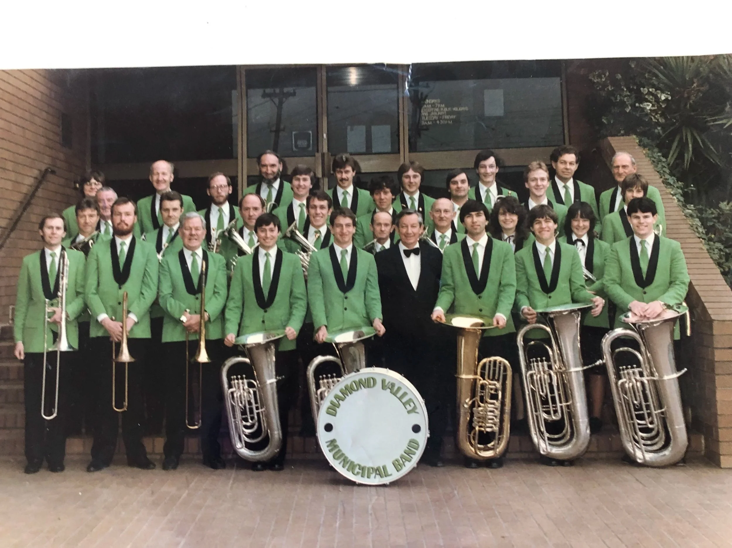 Large group of musicians in matching green jackets, standing outdoors on a brick path, holding brass and percussion instruments, with a sign reading 'Diamond Valley Municipal Band' in front.