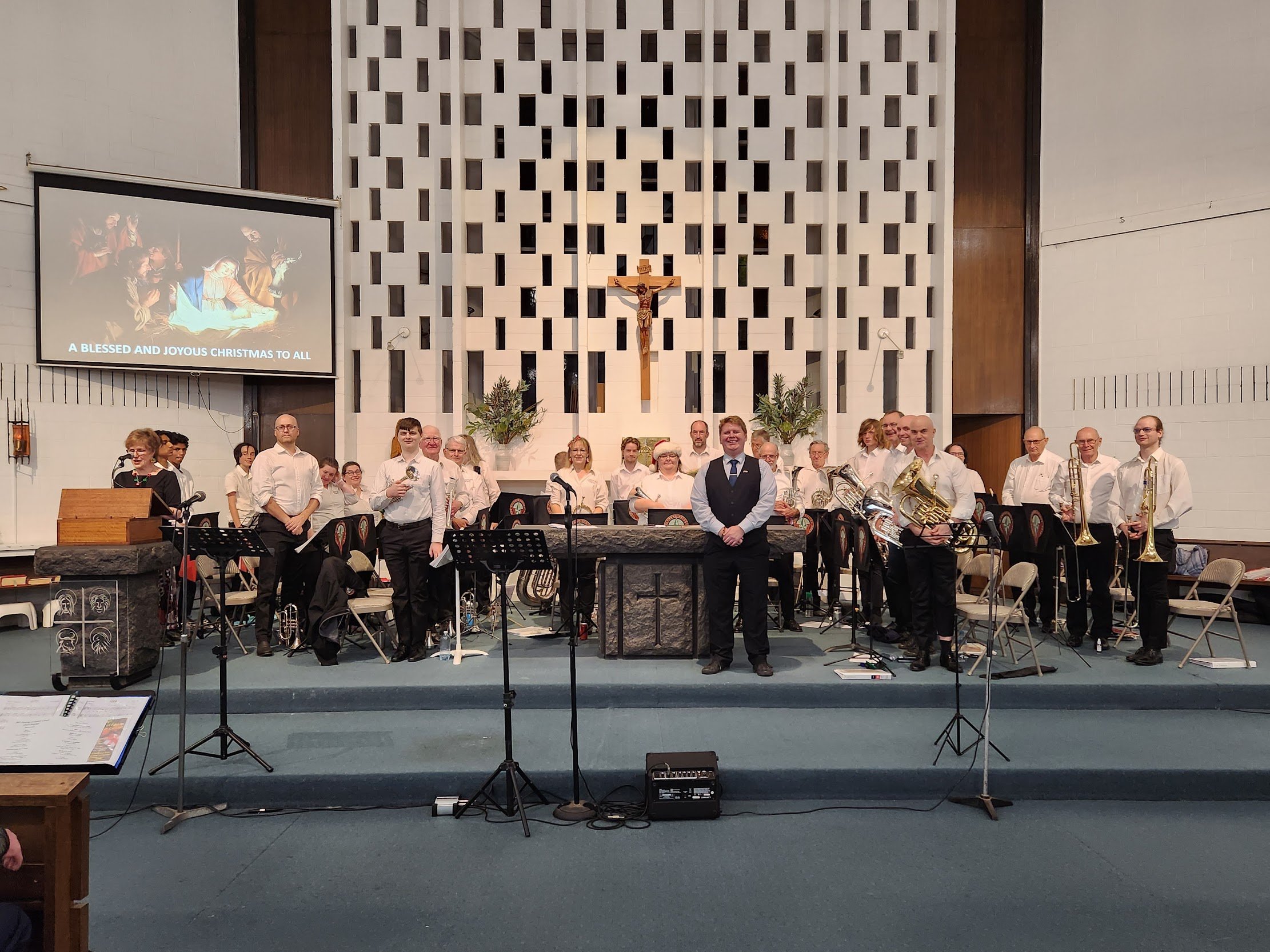 A group of musicians and a conductor standing on a stage inside a church, with a crucifix and decorative wall panels behind them. There is a choir on the left side and music stands in front of the musicians.