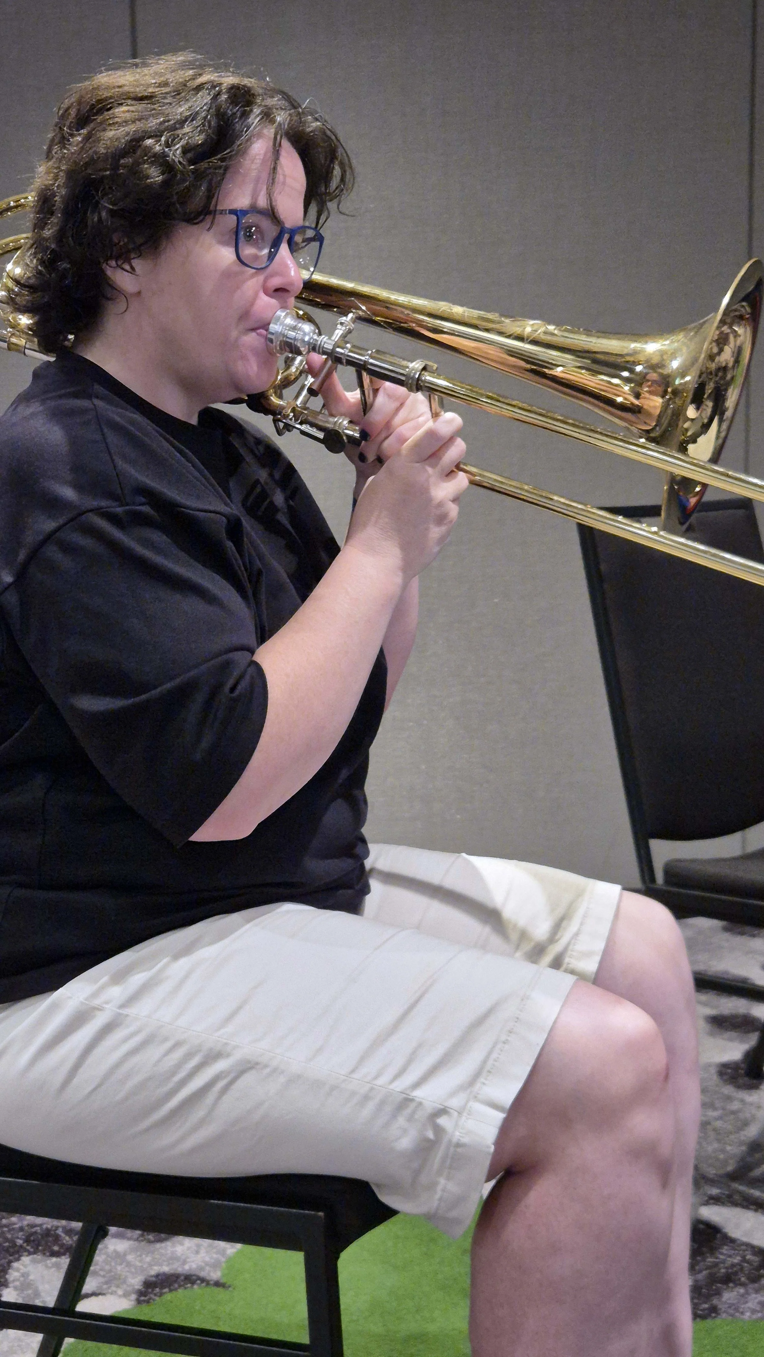A person sitting on a black chair playing a trombone, wearing a black shirt, beige shorts, and glasses.