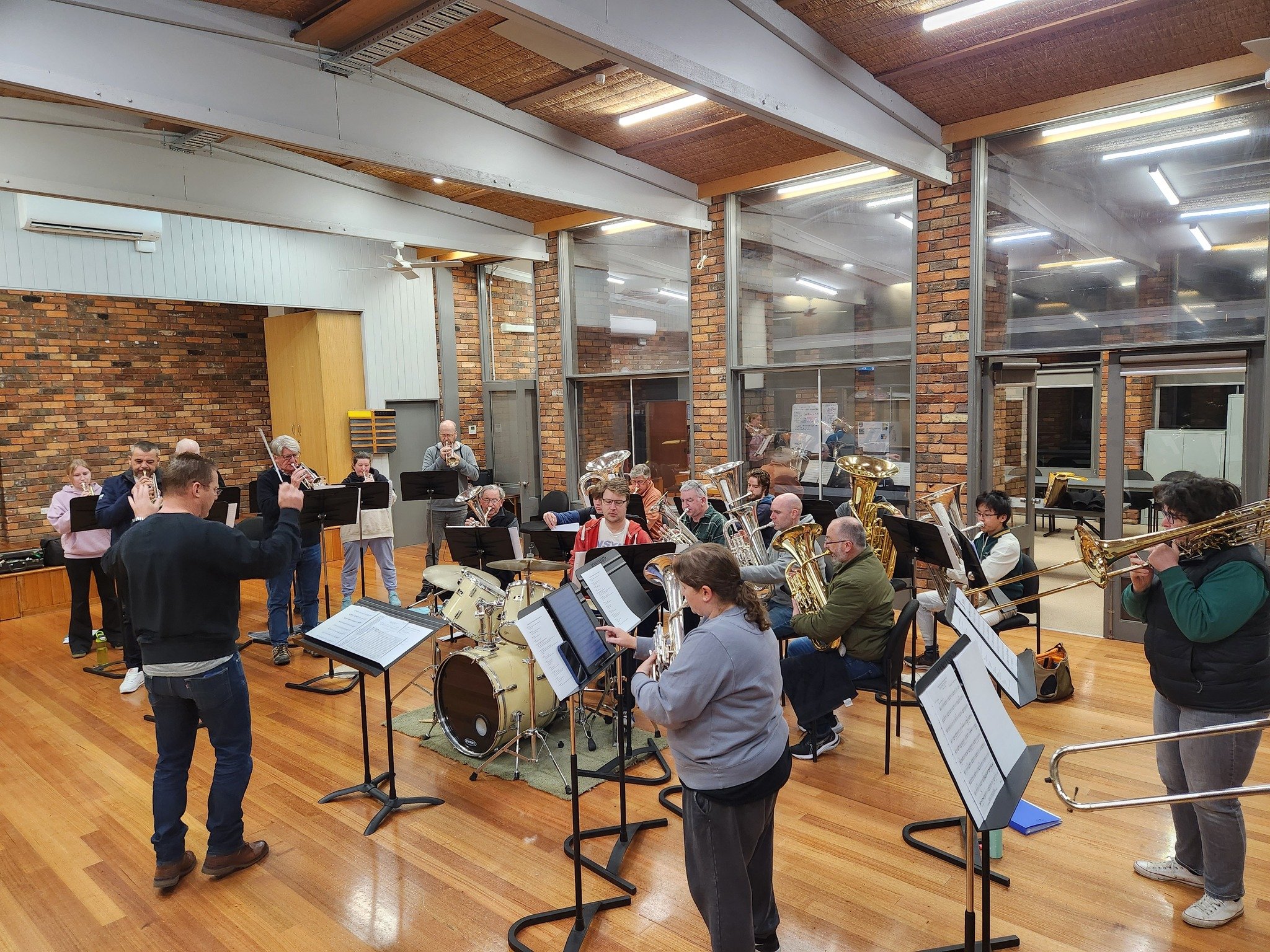 A group of musicians rehearsing in a spacious room with wood flooring, brick walls, large glass windows, and white ceiling beams. The musicians are playing various instruments, including brass, percussion, and woodwinds, with music stands in front of