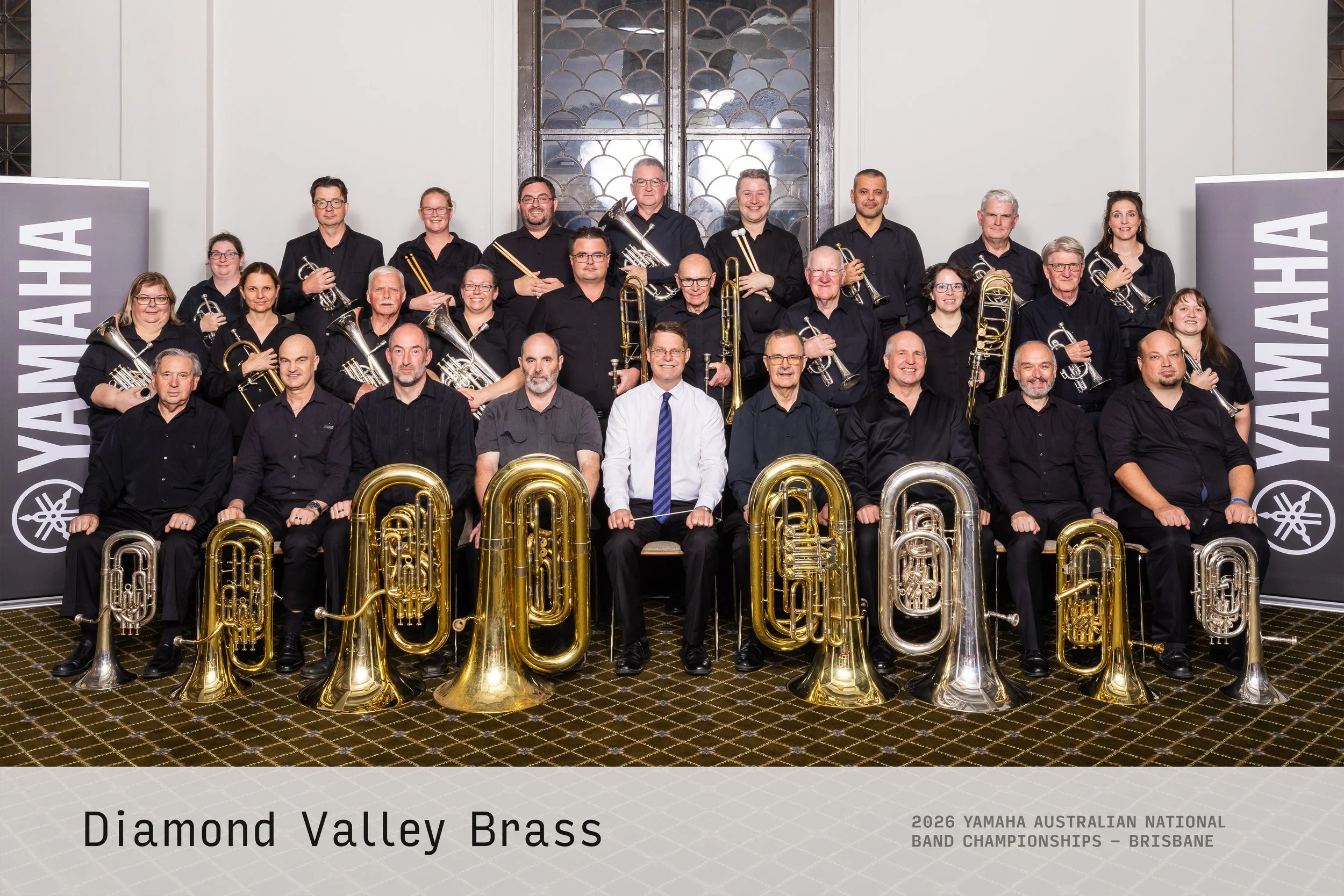 A large group of musicians holding brass instruments, posing for a photo at the 2026 Yamaha Australian National Band Championships in Brisbane. They are in a room with white walls and a decorative window behind them, flanked by Yamaha banners.