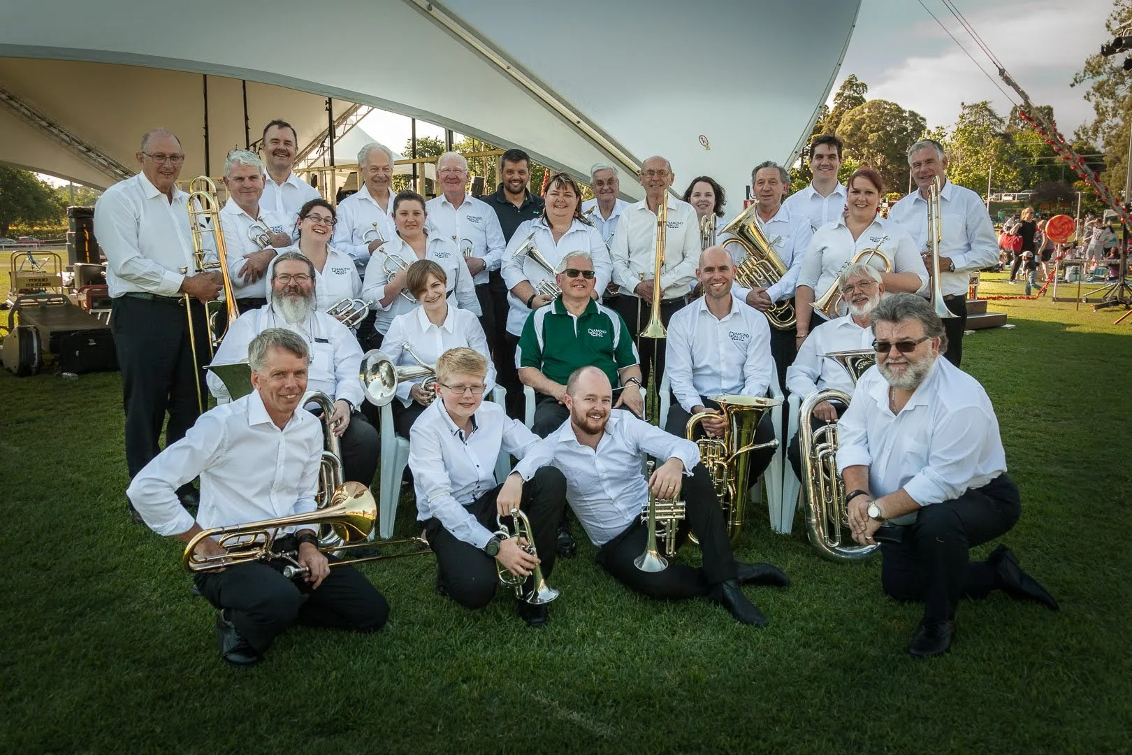 A large group of adults and children posing outdoors with musical instruments, likely a band, on a grassy area under a large tent, with some trees and carnival rides or booths in the background.