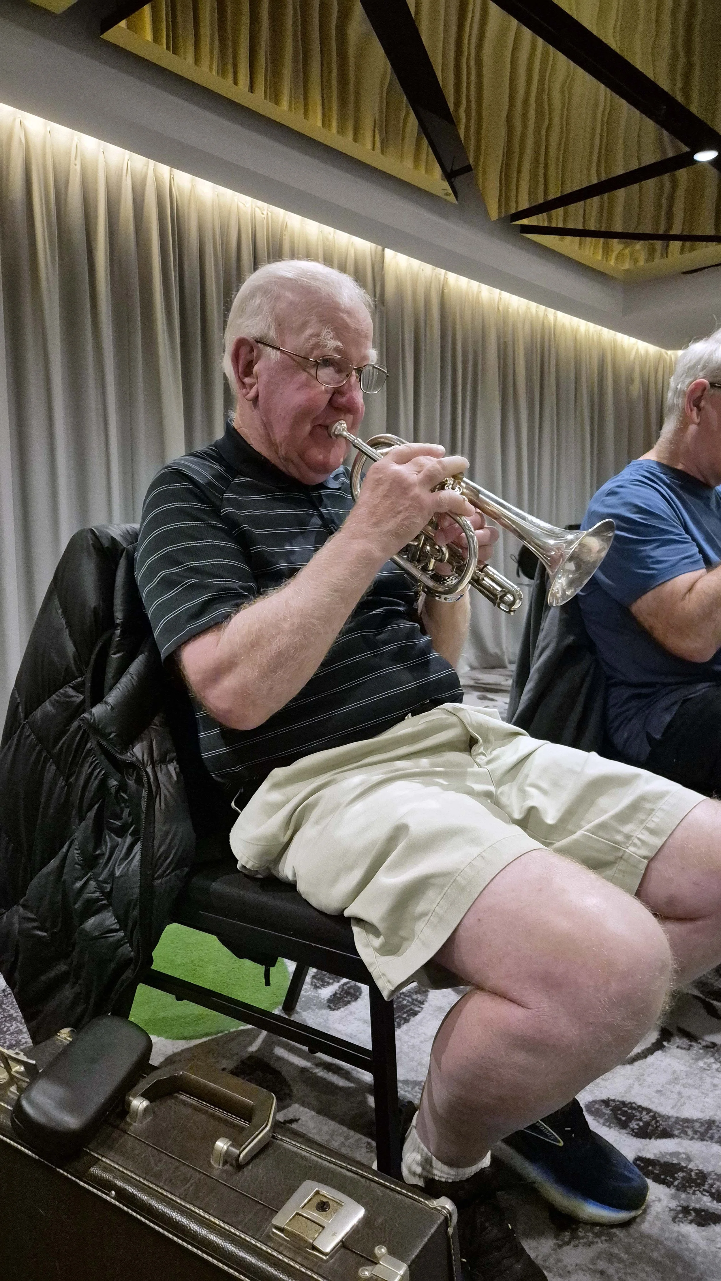An elderly man with glasses wearing a black striped polo shirt and khaki shorts, sitting on a chair, playing a silver trumpet. He has a black jacket draped over the back of his chair. Next to him, another person is partially visible. The setting appears to be indoors with beige curtains and a ceiling with wooden paneling.