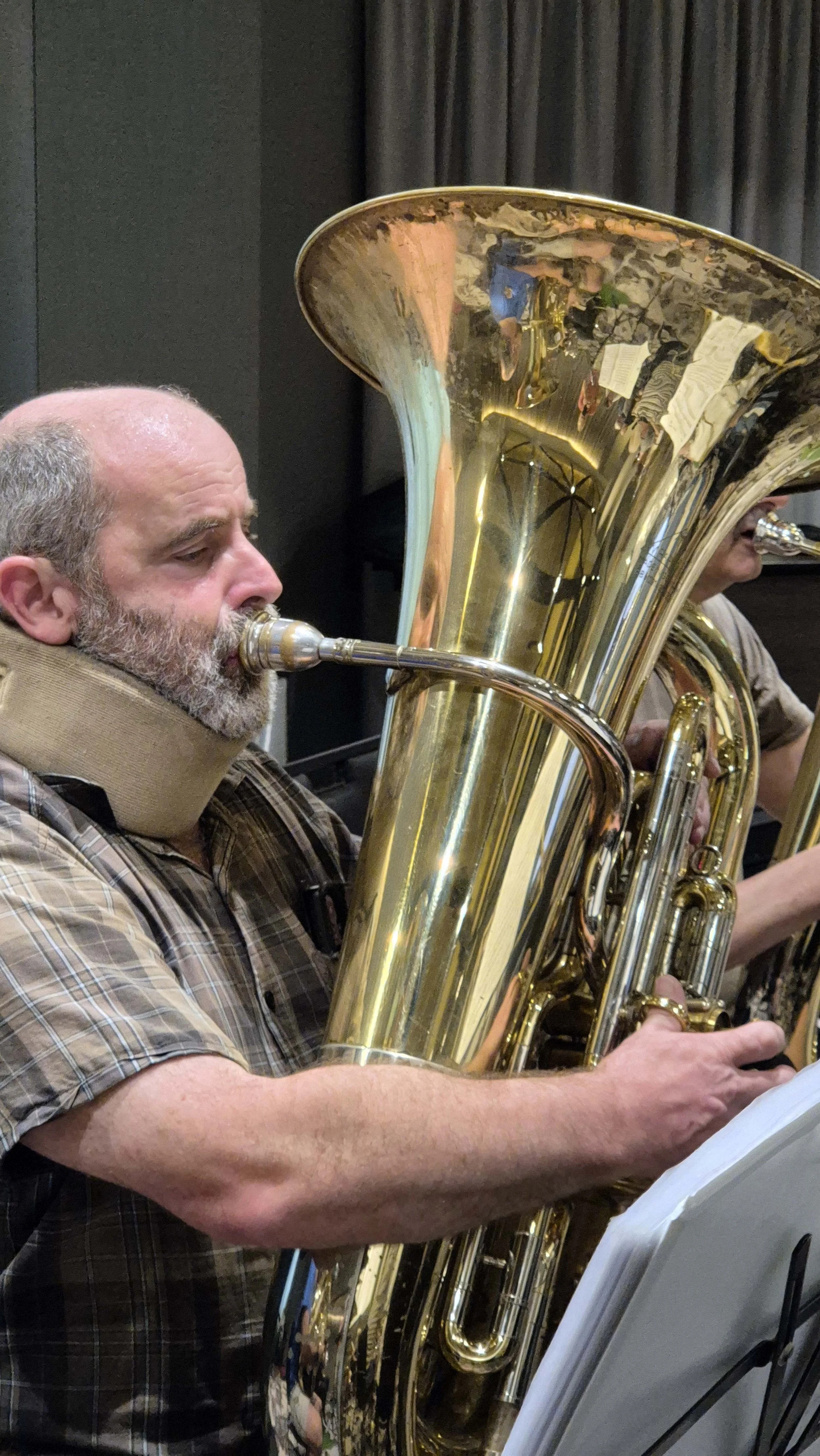 A man with a gray beard playing a baritone saxophone during a musical rehearsal or performance. He is focused, wearing a plaid shirt, and reading sheet music on a stand.