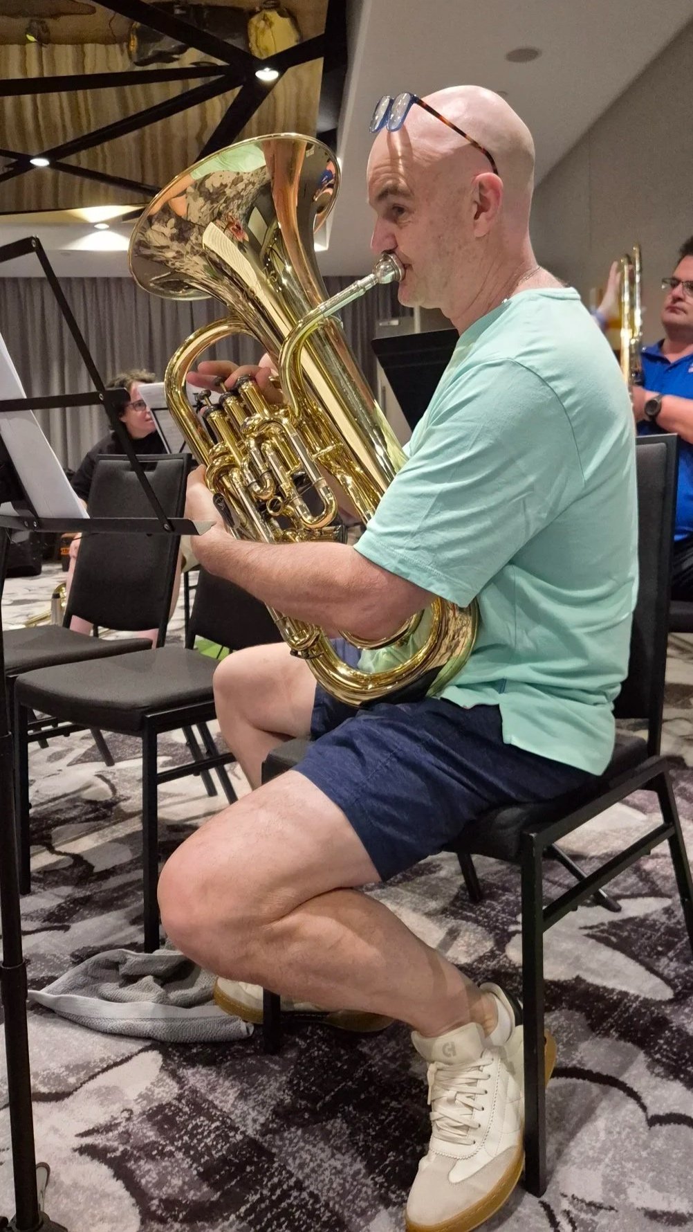 A bald man playing a brass euphonium during an orchestra rehearsal or performance. He is wearing glasses on his head, a light green t-shirt, dark shorts, and white sneakers. Other musicians and sheet music stands are visible in the background.