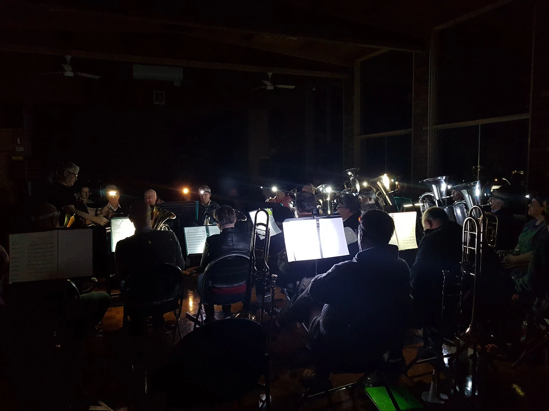 An indoor concert or performance with a group of musicians, including wind instrument players, illuminated by stage lights in a dark room, with audience seated in front.