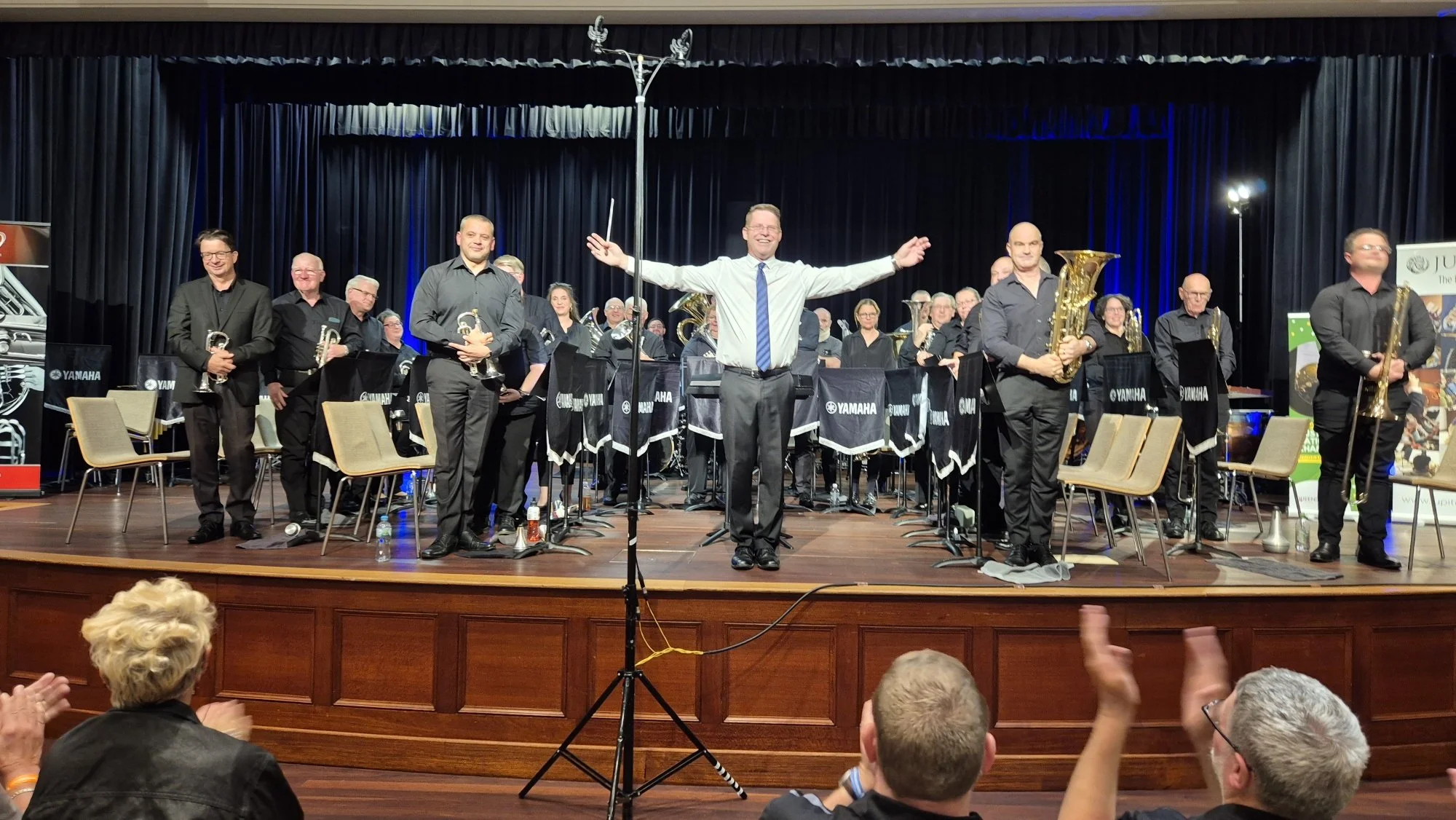 A large group of musicians on stage, with a conductor at the center raising his arms, in front of a black curtain backdrop. The musicians are holding brass instruments like saxophones and trumpets, wearing black attire, and there are chairs and music