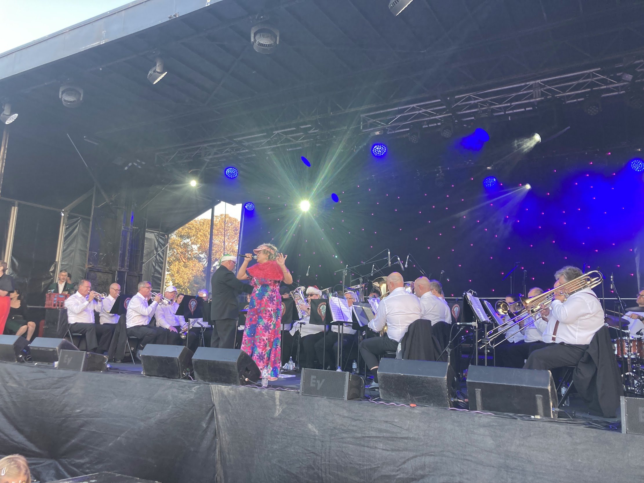 Brass Band  performing on an outdoor stage with a female conductor wearing a colorful floral dress, and a large group of musicians playing instruments, under stage lighting and a partially open black canopy.