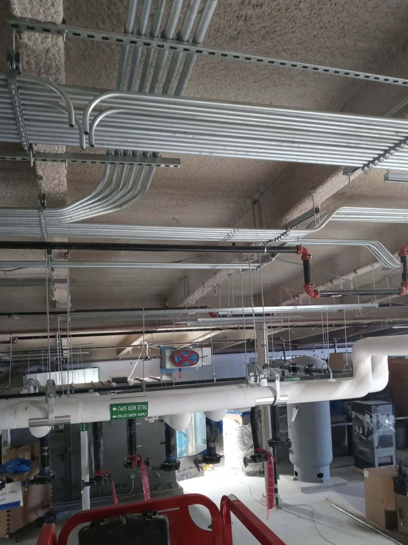 Ceiling with silver metallic pipes and conduit, some with red valves, in a mechanical or utility room.