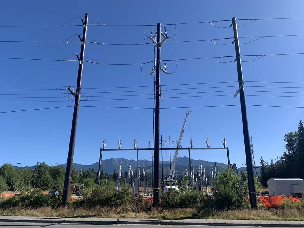 Power lines and electrical poles at a power substation with a crane working on infrastructure, mountains in the background and clear blue sky.