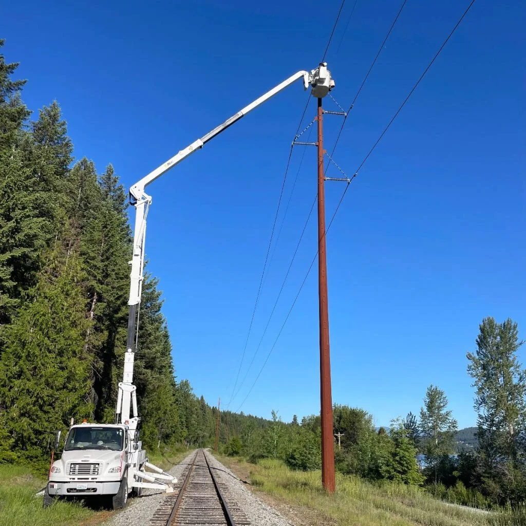 A utility worker in a bucket lift is repairing power lines on a tall wooden utility pole beside a railroad track in a rural area with trees and a blue sky.
