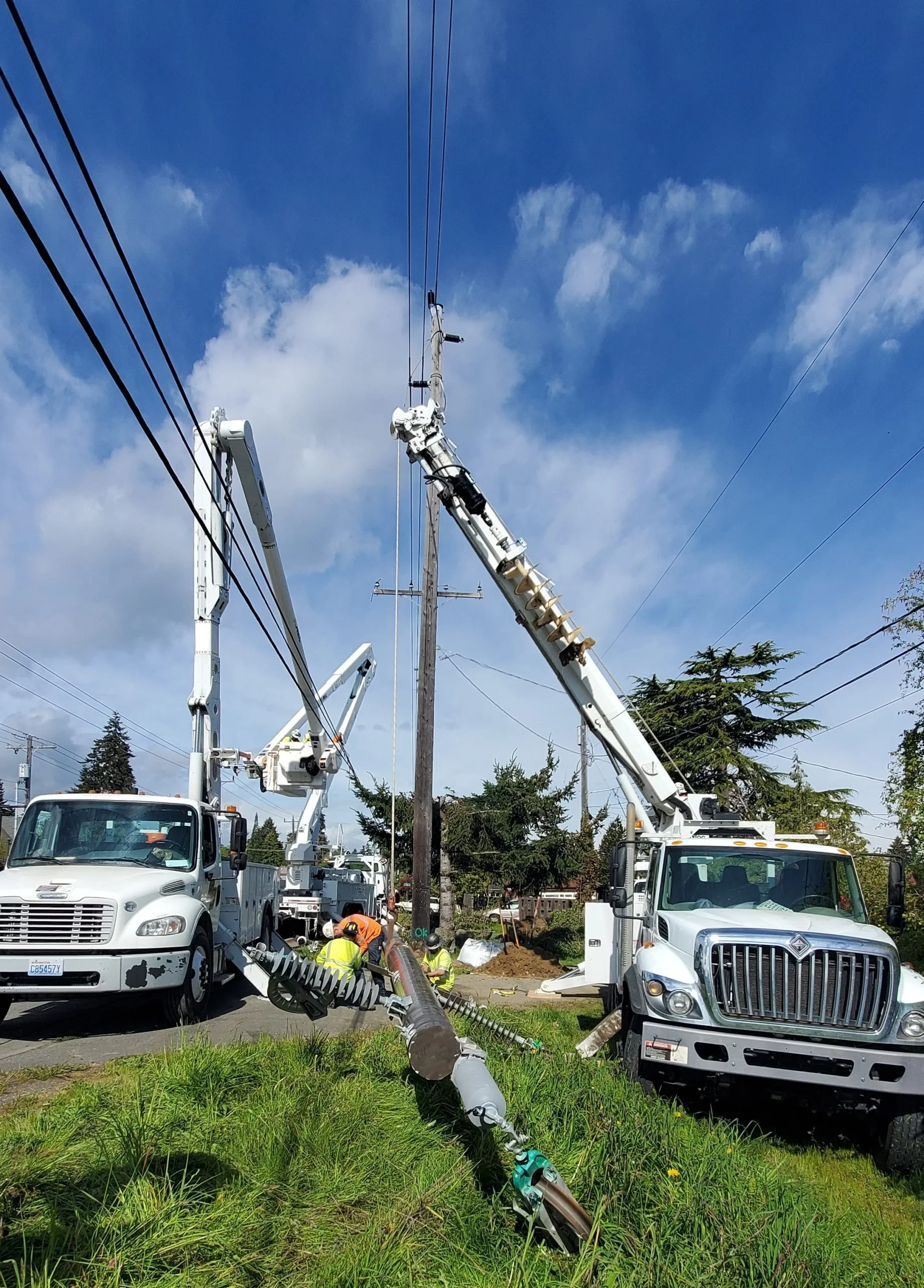 Workers repairing a toppled utility pole with power lines using bucket trucks on a roadside with cars parked nearby, trees, and a partly cloudy sky.
