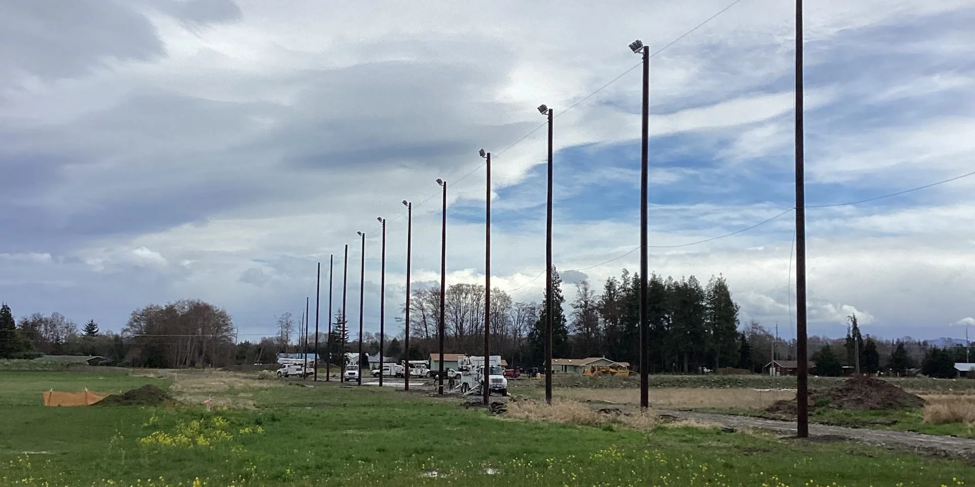 A row of tall wooden light poles with attached floodlights stretches into the distance in an open field. Several vehicles, including trucks, are parked near small houses or buildings in the background, with trees and a cloudy sky overhead.