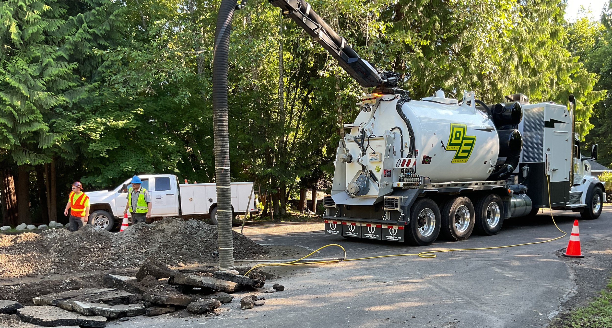 Workers in safety vests and helmets inspecting a construction site with a large vacuum truck cleaning a utility pole on the street, surrounded by green trees and construction cones.