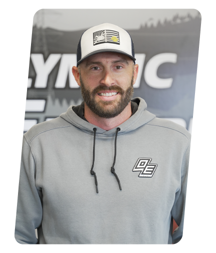 A man with a beard wearing a gray hoodie and a white cap with an American flag and yellow stripe, smiling in front of a black and white background with the words 'SWYOGIC'.
