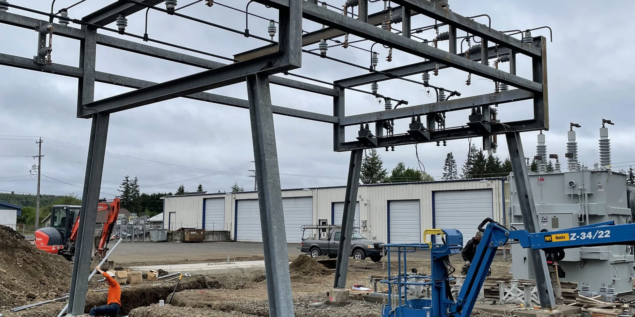Construction site with electrical infrastructure being installed, workers, construction equipment, and storage units under an overcast sky.