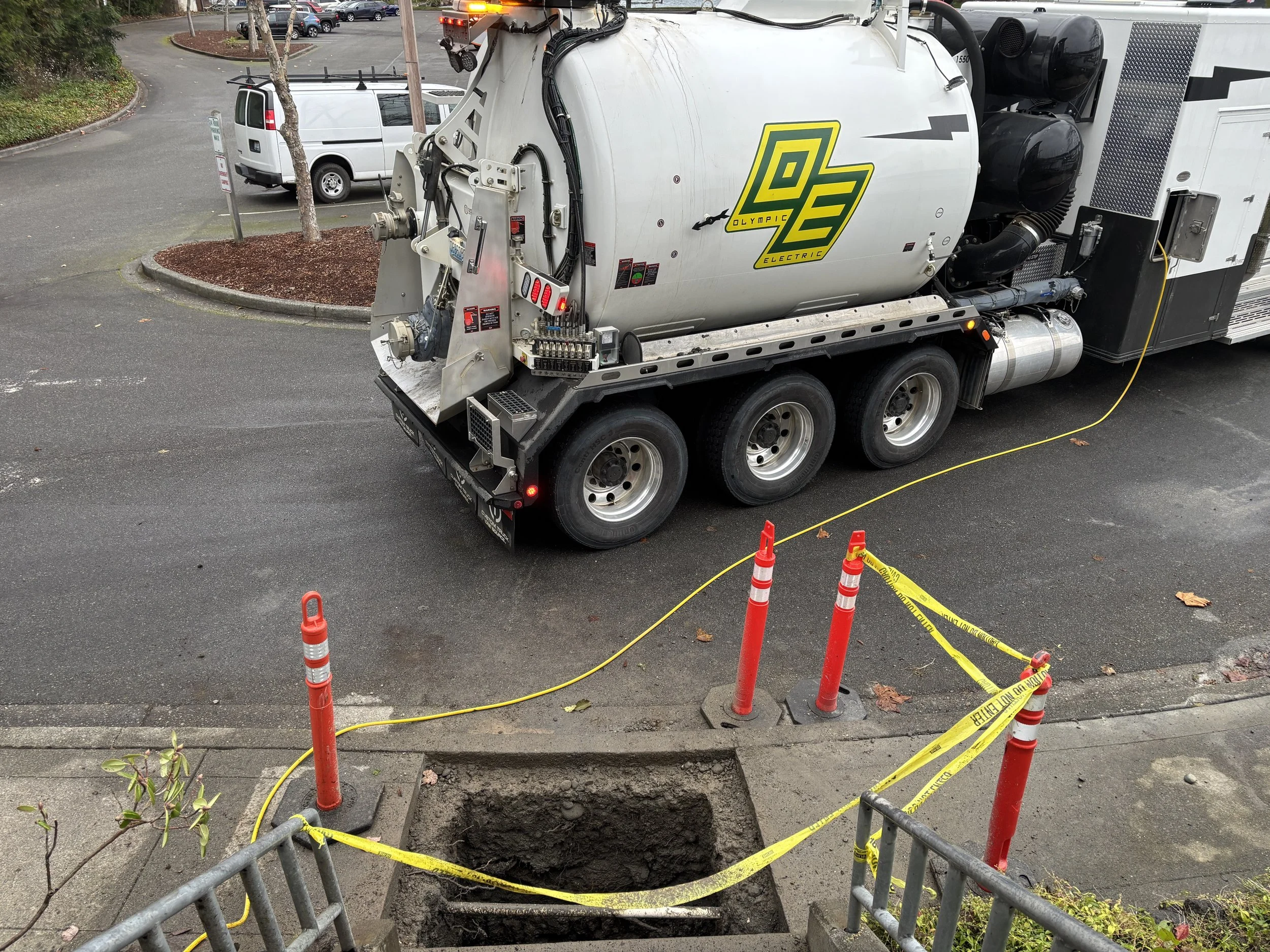 An open manhole on a city sidewalk surrounded by red and yellow caution cones and tape. In the background, there is a large white street cleaning truck and a white van parked nearby. Two small trees are planted in circular mulched areas.