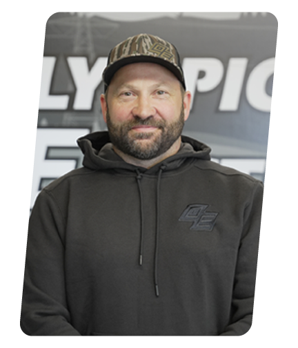 A man with a beard wearing a black hoodie and camouflage baseball cap standing in front of a backdrop with the Olympic logo.