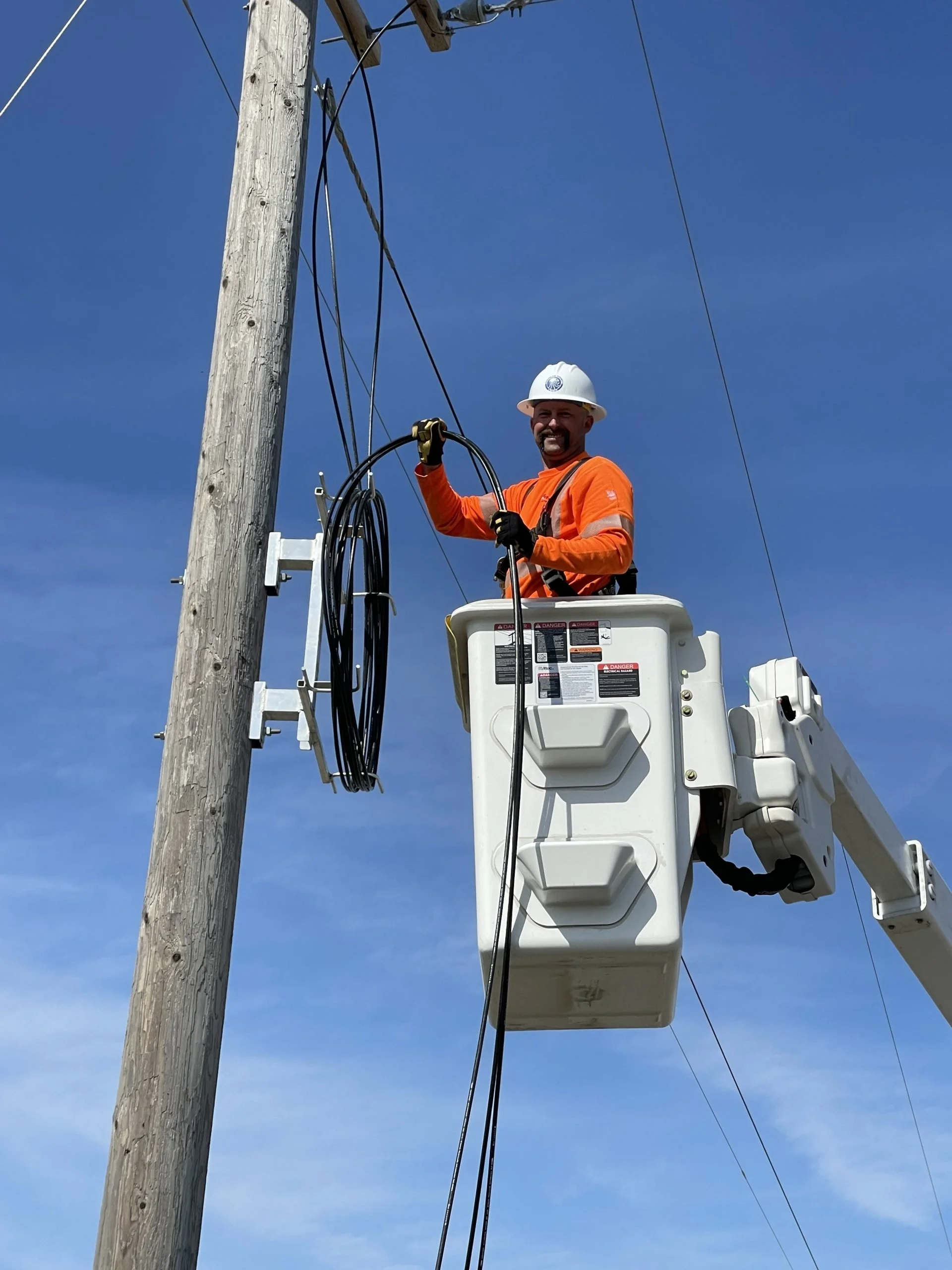 A utility worker wearing an orange safety shirt, a white helmet, and gloves, standing in an elevated bucket lift working on power lines next to a wooden utility pole against a clear blue sky.