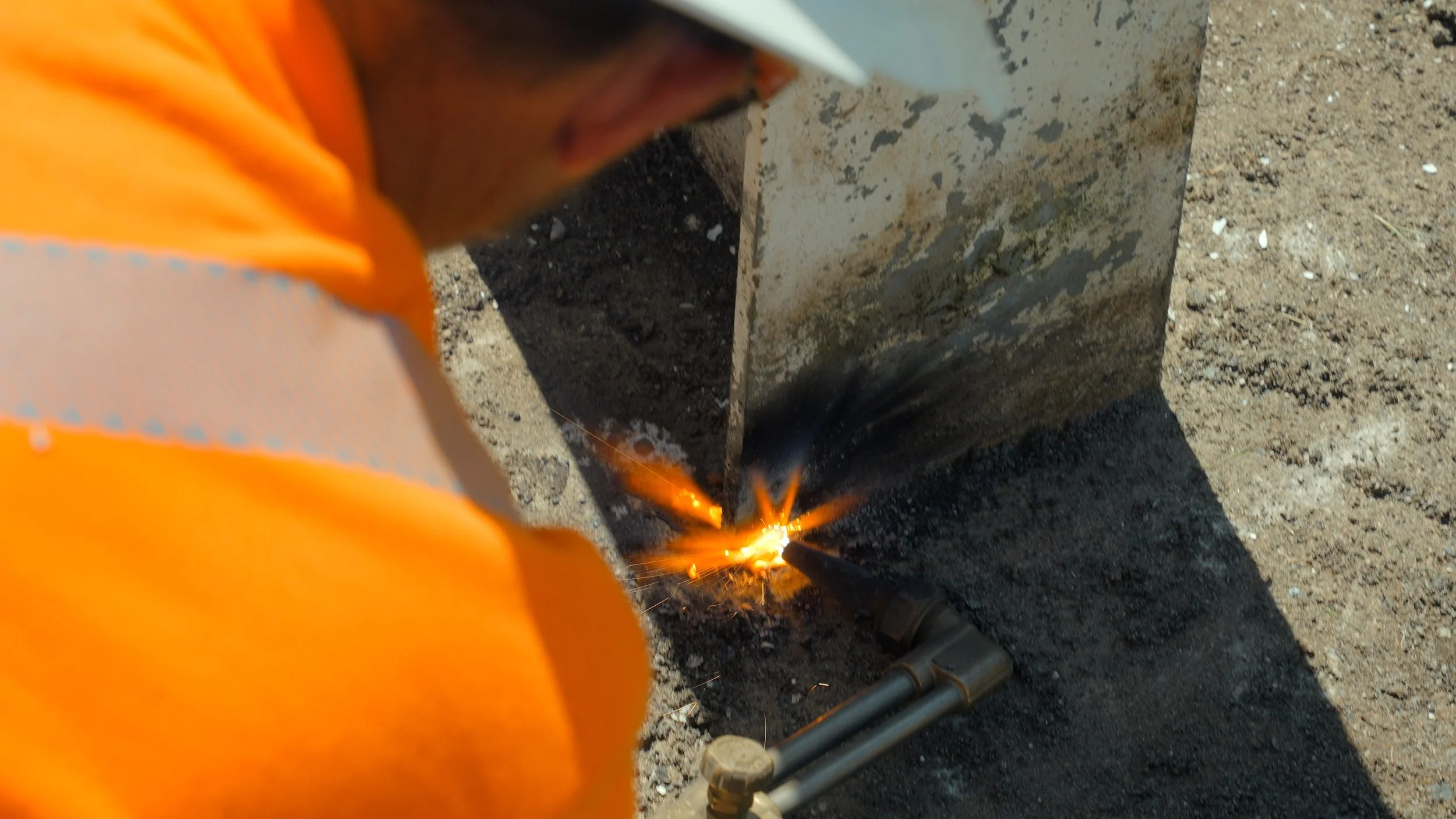 A construction worker in an orange jacket and white helmet welding metal on the ground outdoors, with sparks flying from the welding process.