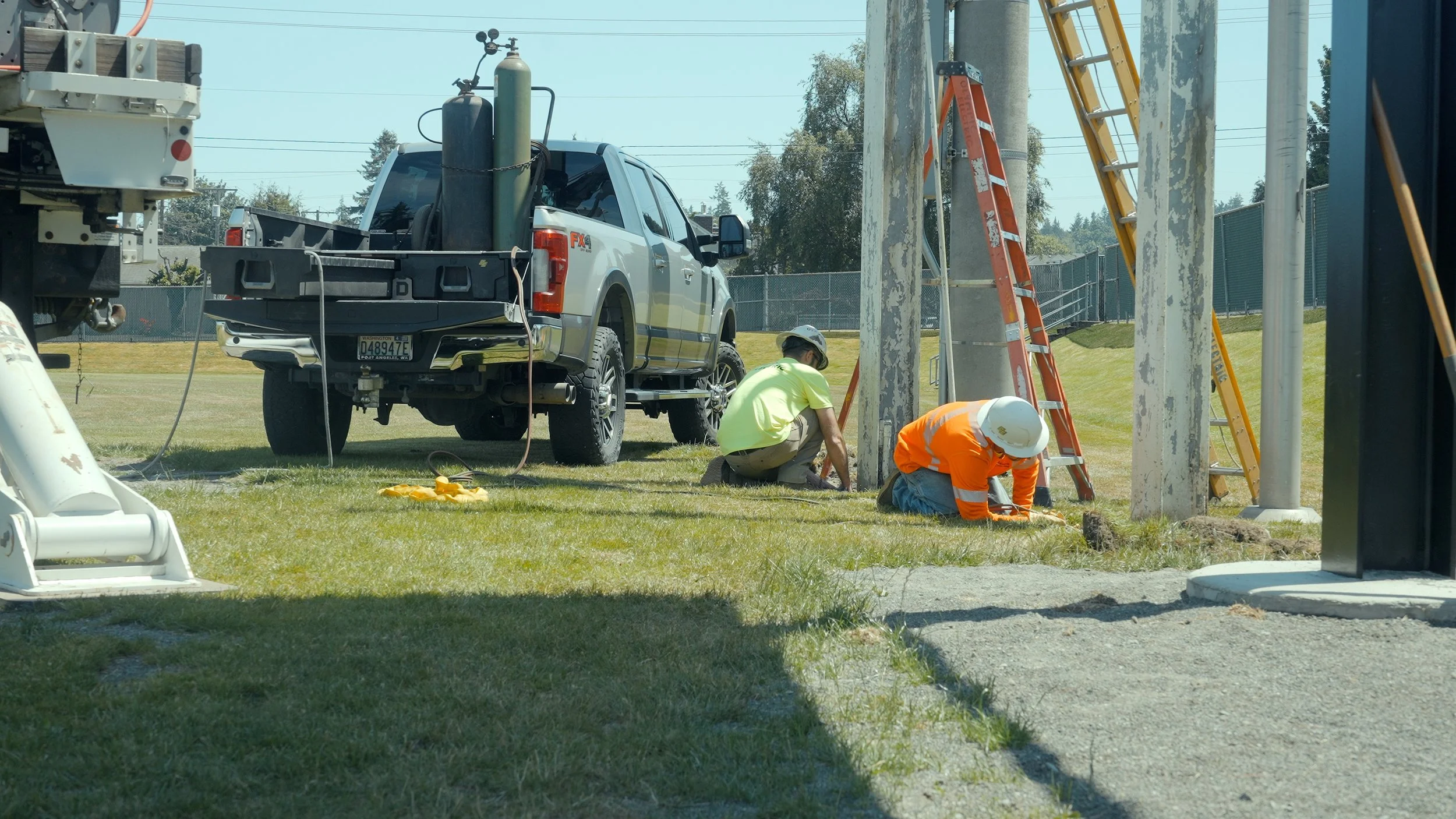 Two construction workers wearing safety helmets working on an outdoor electrical pole, with a pickup truck parked nearby and ladders leaning against the pole.