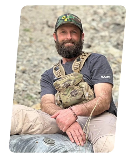 A man with a beard smiling, sitting outdoors on rocky terrain, wearing a camouflage cap with 'OEF' and 'OEF Elite' written on it, a gray T-shirt, beige pants, and a camouflage chest rig.