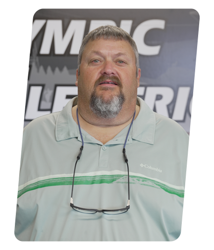 A man with short hair and a beard wearing a light-colored polo shirt and glasses hanging around his neck. He is standing in front of a background with partially visible text.