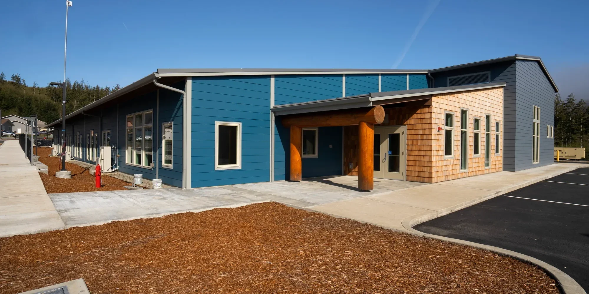Modern two-story building with blue and natural wood siding, multiple windows, and a covered entrance supported by large wooden logs. Surrounding parking lot and landscaped area with mulch.