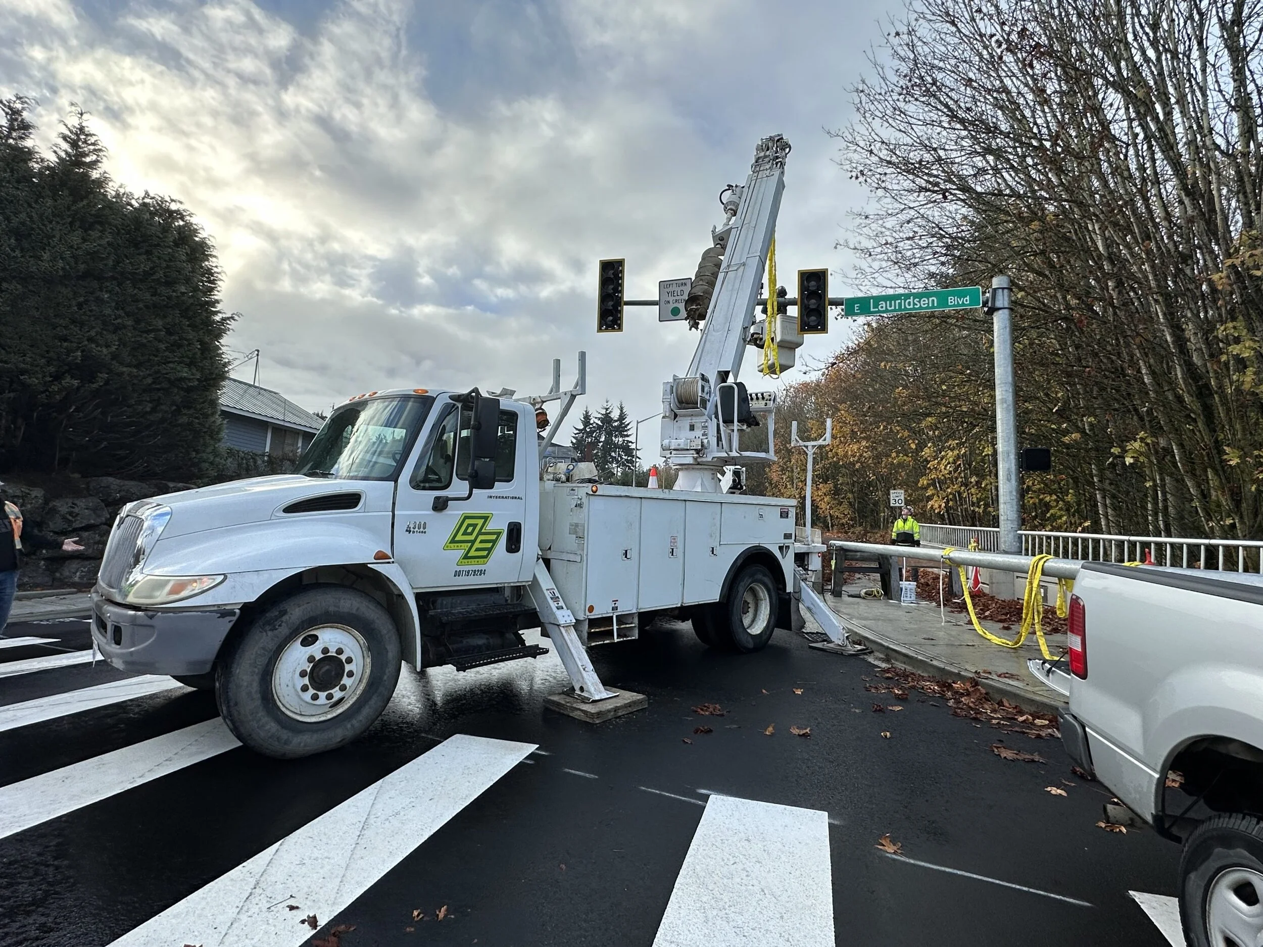 Utility bucket truck with a cherry picker lift positioned at a traffic light in an urban area. The truck is white with green company logo, parked on a crosswalk. There is a street sign for E Lauridsen Blvd, traffic light, and a worker in a yellow safety vest nearby. Trees with autumn leaves and a house are in the background, under a cloudy sky.