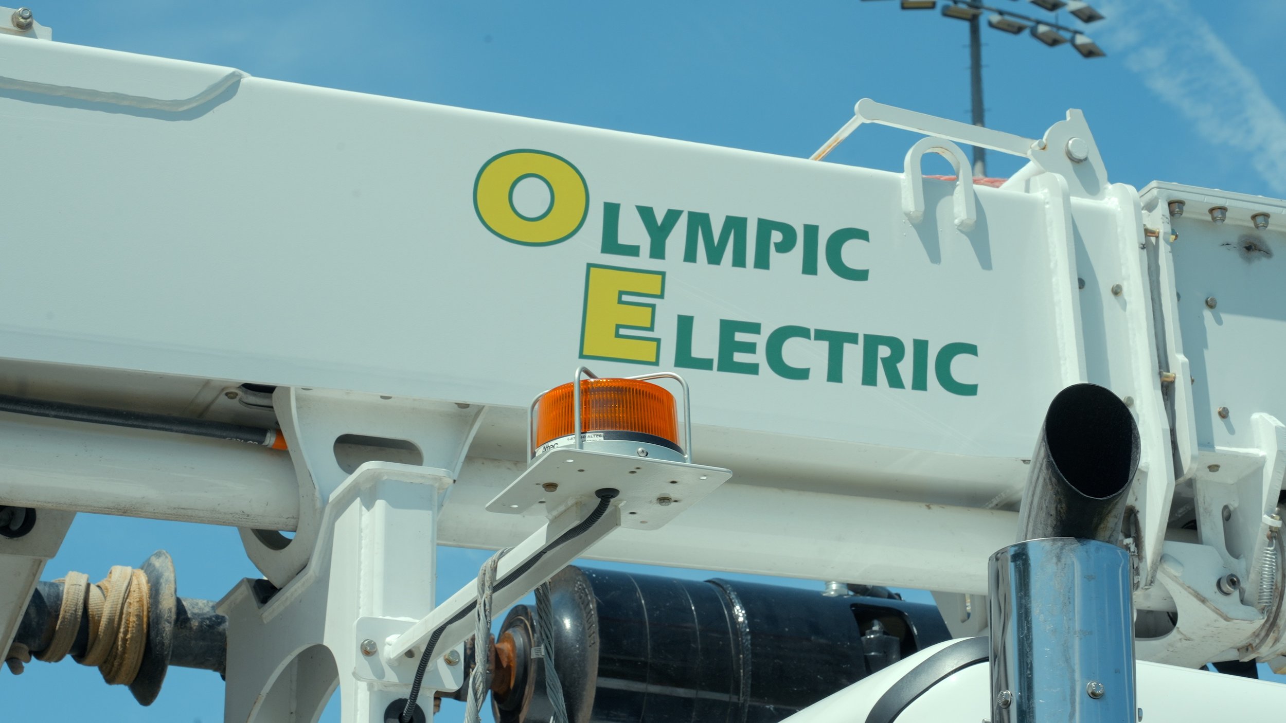 Close-up view of a white electric utility truck with a logo reading 'Olympic Electric' on the side, under a blue sky with some clouds. The truck has orange warning lights and various mechanical parts visible.