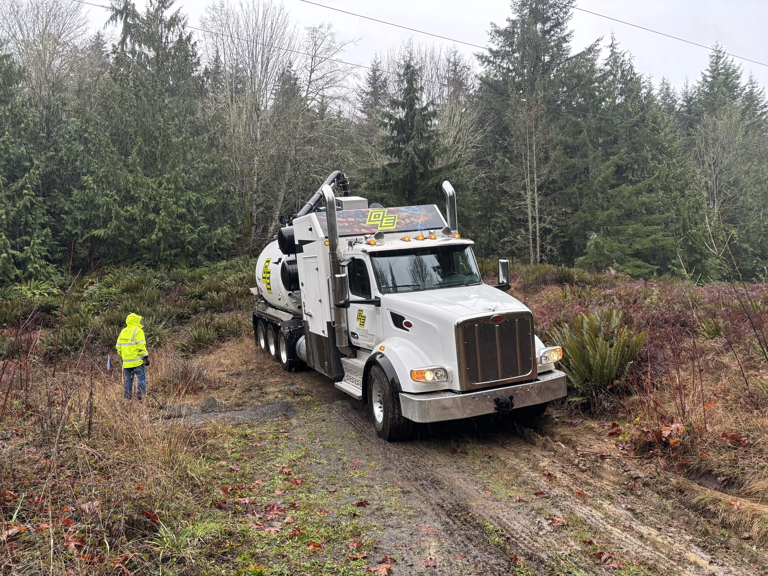 A large white utility truck with equipment on top is parked on a muddy forest trail, with two workers in yellow high-visibility jackets nearby and trees in the background.