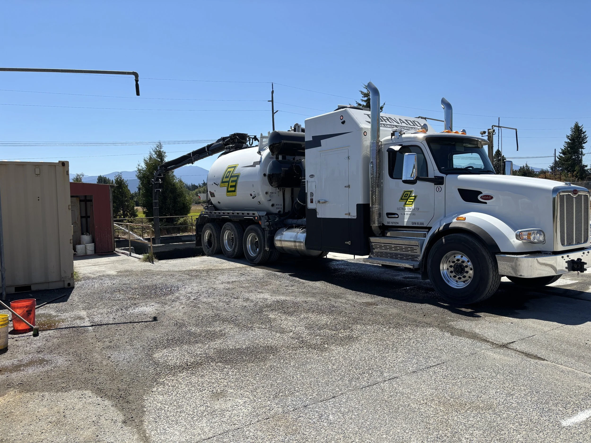 A large white truck with a black arm and logo on the side parked on gravel ground, with trees, utility poles, and mountains in the background under a clear blue sky.