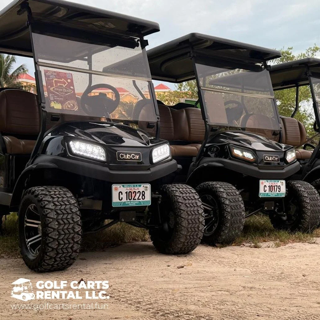 Two black golf carts with brown seats parked on a grassy area. Each cart has a clear windshield, large tires, and a Belize license plate. The carts are branded with Club Car. The background shows trees, buildings, and a cloudy sky.