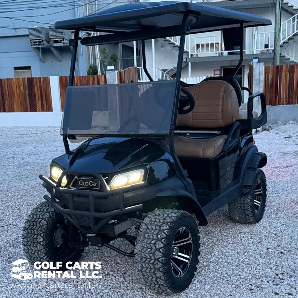 Black golf cart with tan seats and a roof, parked on gravel, with a wooden fence and residential building in the background.