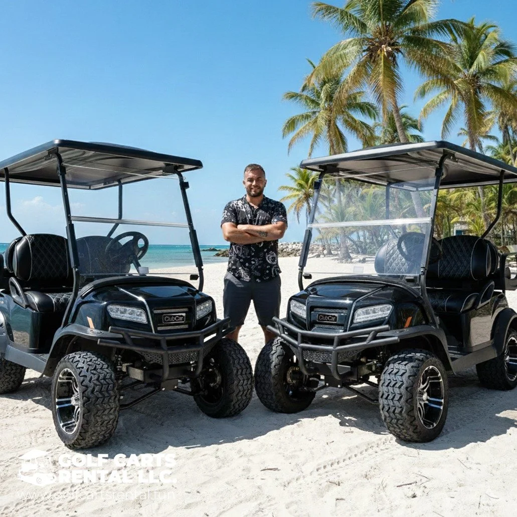 A man standing on a beach between two black golf carts with palm trees and the ocean in the background.