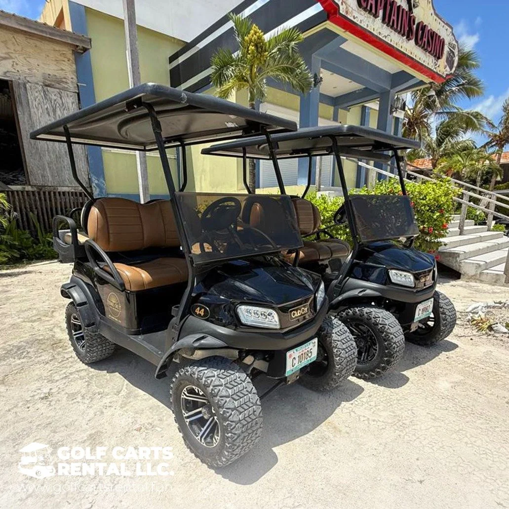 Two black electric golf carts parked side by side on a sandy surface in front of a building with a sign that reads 'CaptaIns Casino'. Each cart has brown seats, a windshield, and a roof. The background includes palm trees, stairs, and a cloudy sky.