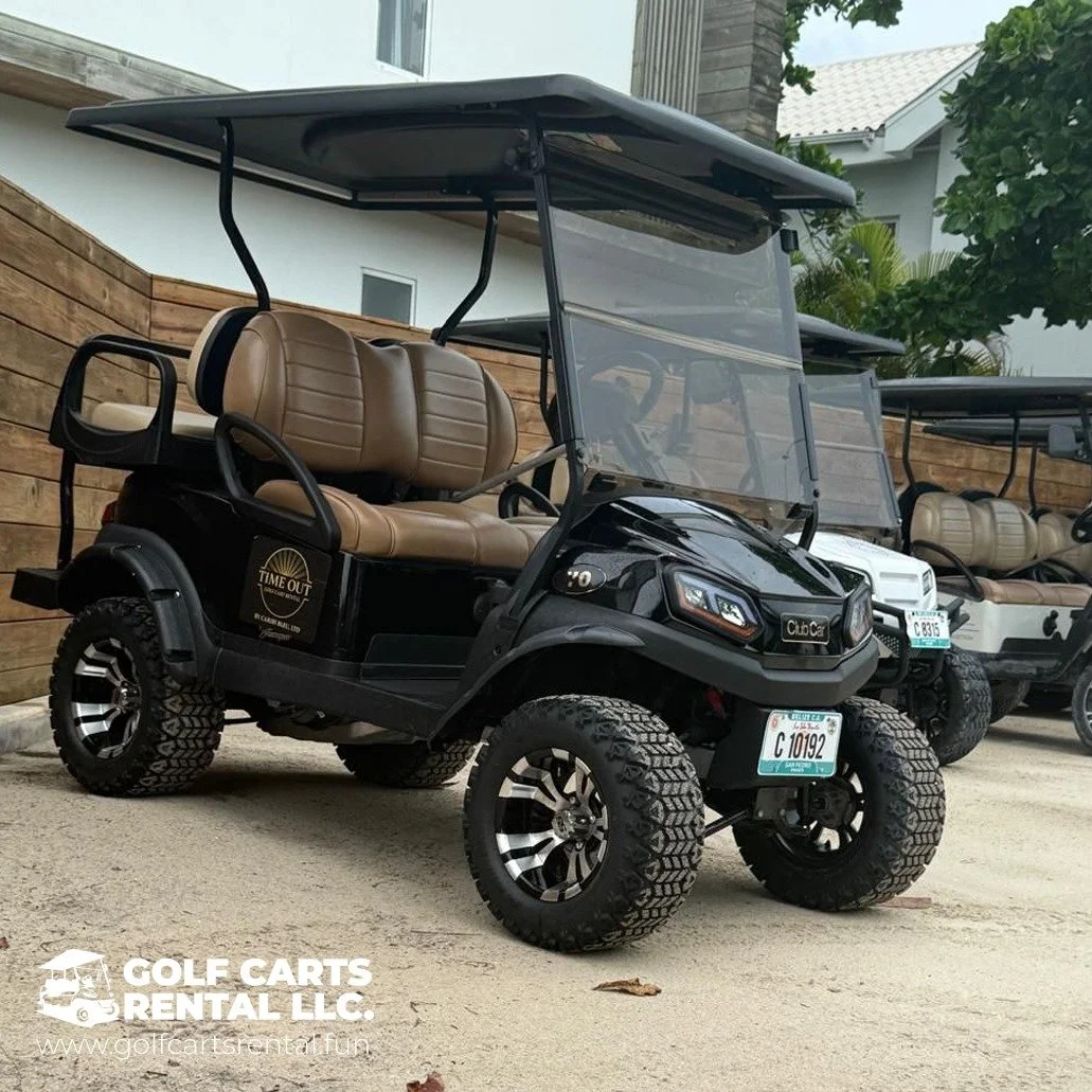 A black and tan golf cart with off-road tires parked on a sandy surface. The golf cart has a tan back seat, a windshield, and a roof, with additional golf carts visible in the background. There is a wooden fence and a modern building behind it, with 