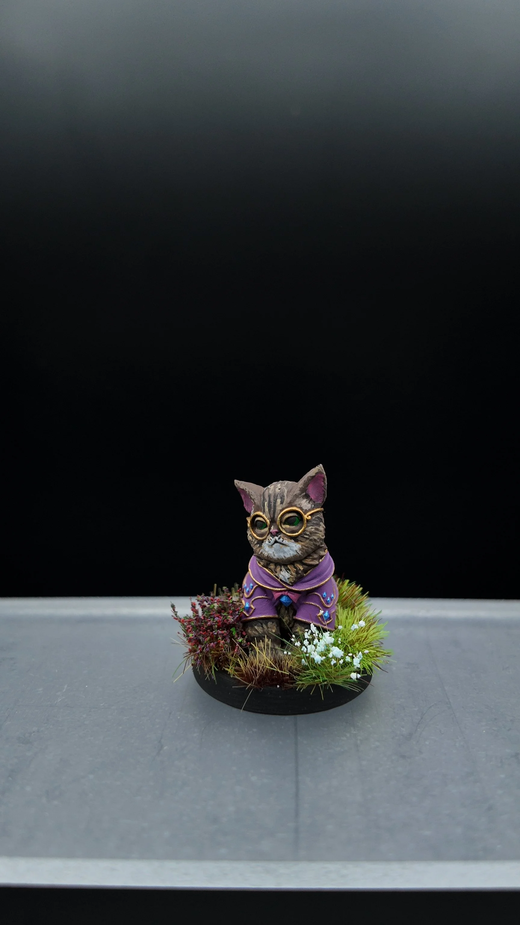A small cat figurine dressed in purple robes with glasses, surrounded by plants with red and white flowers, against a black background.