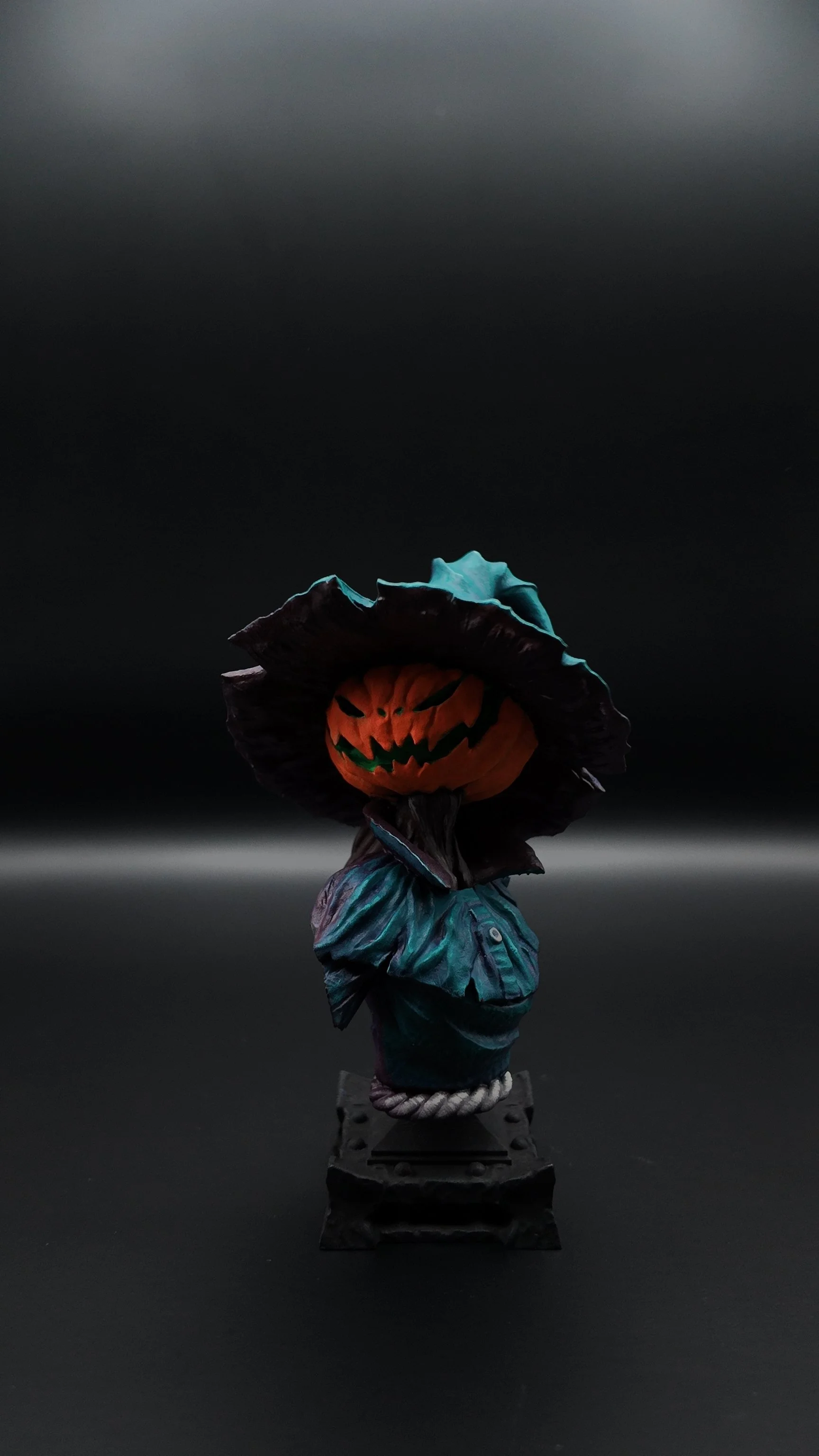 A Halloween-themed figurine with a pumpkin head, carved to have a menacing face, wearing a black wide-brimmed hat and a blue-green jacket, standing on a small pedestal against a dark background.