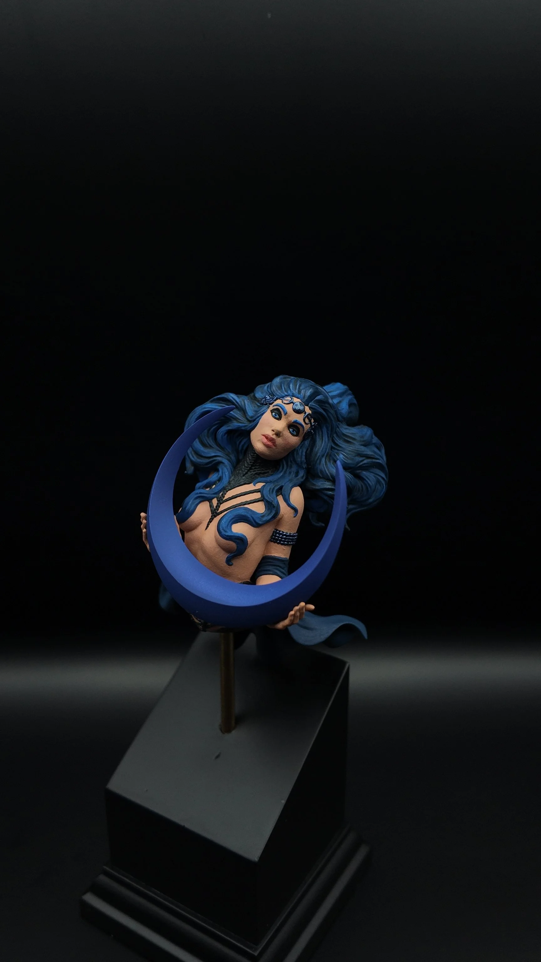 A detailed sculpture of a woman with flowing blue hair, holding a blue crescent-shaped object, on a black display stand against a dark background.