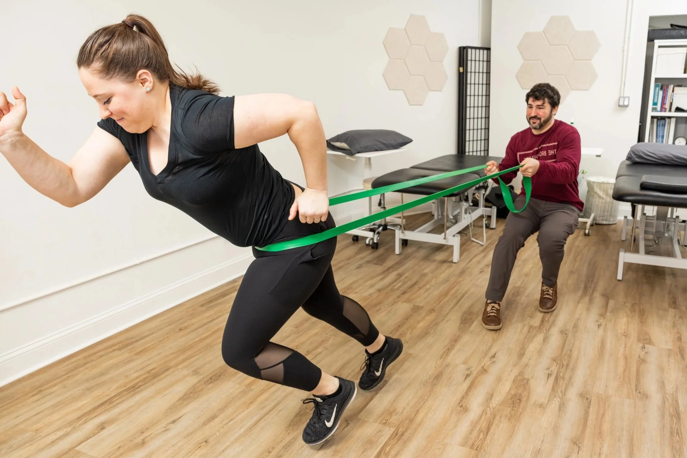 A woman participating in physical therapy exercises with assistance from a man holding resistance bands. The woman is bending forward, and the man is kneeling, providing support during the exercise in a clinical setting with treatment tables and medical equipment.