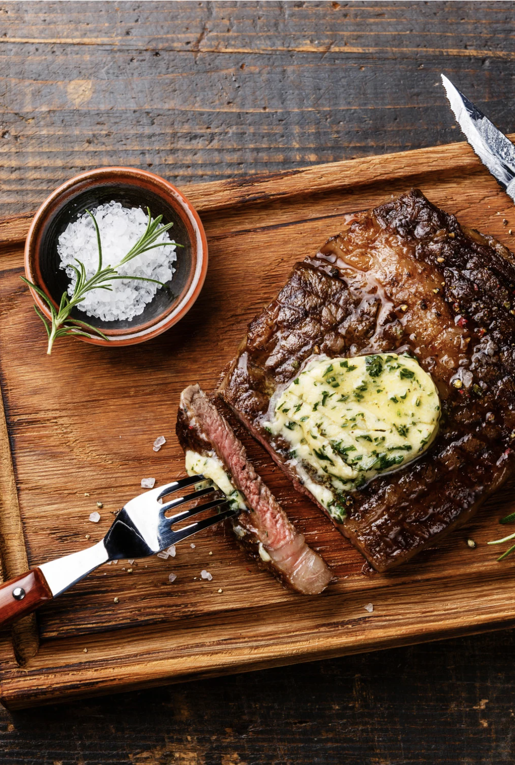 A wooden board holds a cooked steak topped with herb butter, with a piece cut and on a fork nearby. A small bowl of coarse salt and a sprig of rosemary are also on the board. A knife is visible at the edge of the board.